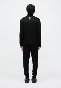 Μαύρο φόρμα που αποτελείται από ζακέτα με φερμουάρ και στενές παντελόνια. Διαθέτει ψηλό γιακά, ριμπ μανσέτες και ένα μίνιμαλ ριγέ διακοσμητικό στο πίσω μέρος.