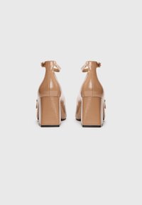 Béžové lakované kožené boty na blokovém podpatku s kotníkovými pásky, s texturovaným povrchem a elegantním, moderním designem.