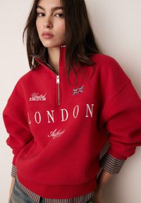 Felpa rossa con scritte bianche "LONDON" e "United Kingdom." Presenta un colletto con mezza cerniera e polsini a righe, realizzata in materiale morbido.