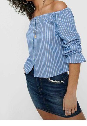 Kobieta w niebiesko-białej bluzce odsłaniającej ramiona w paski z marszczonymi rękawami oraz ciemnoniebieską mini spódniczką dżinsową z koronkowym wykończeniem na kieszeni.