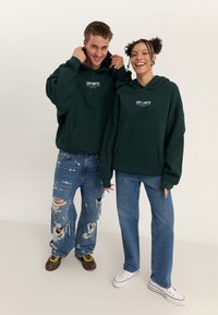 Green hooded sweatshirt with “OFF LIMITS” text, paired with distressed denim jeans. Casual and relaxed fit with front pocket detail.