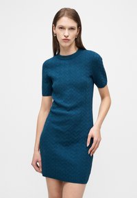 Γυναίκα με ίσια καστανά μαλλιά που φοράει υφασμάτινο φόρεμα κοντών μανικιών σε τυρκουάζ μπλε και χρυσούς κρίκους σκουλαρίκια, στέκεται μπροστά σε λευκό φόντο.