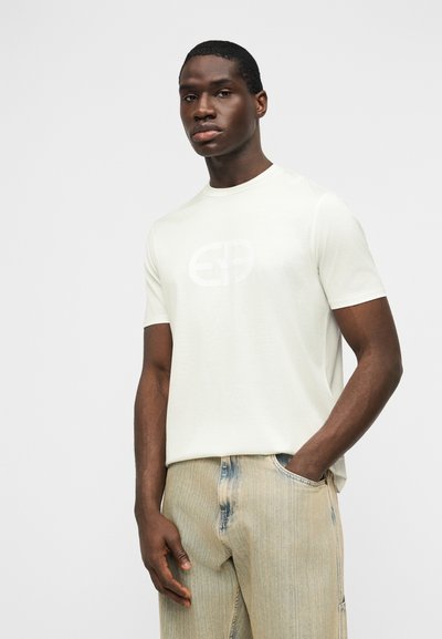 Άνδρας που φοράει λευκό μπλουζάκι με κοντά μανίκια και διακριτικό λογότυπο, καθώς και ανοιχτόχρωμο ξεθωριασμένο τζιν, στέκεται με το ένα χέρι στην τσέπη.