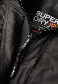 Wnętrze czarnej kurtki z podwójnymi zamkami błyskawicznymi i logiem "SUPERDRY", podszewka z fakturowanej tkaniny, prezentujące elegancki design i funkcjonalne elementy.