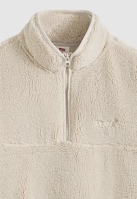 Φλις επαναφοράς σε κρεμ χρώμα με ψηλό γιακά, μισό φερμουάρ και υφή απαλή, εμπλουτισμένη. Διαθέτει διακριτικό λογότυπο στο κάτω μέρος.