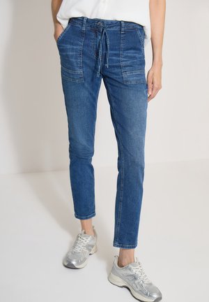 Kvinde iført blå denim joggerbukser med forreste patch-lommer og grå atletiksneakers, står mod en ensfarvet lys baggrund.