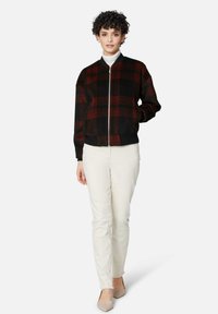 Zwarte en rode geruite bomberjack met geribbelde manchetten en zoom, met sluitclip aan de voorkant, gedragen over een witte coltrui, gecombineerd met crèmekleurige broek.