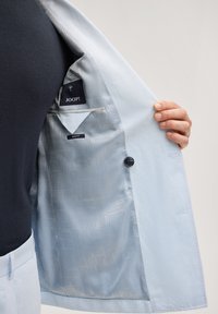 Veste slim fit bleu clair ouverte pour montrer la doublure à motifs et la poche intérieure avec l'étiquette JOOP!, portée sur une chemise foncée.