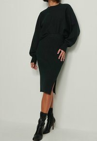 Nainen, joka on pukeutunut mustaan ribattuun midi-hameeseen, jossa on sivupilkku, yhteensopivaan löysään mustaan neuleeseen ja mustiin korkokenkiin, seisoo tasaisella seinällä.