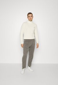 Crème kabelgebreide coltrui, grijze corduroy broek en witte sneakers met groene accenten. Model staat op een effen achtergrond.