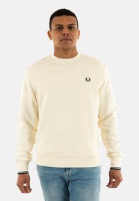 Felpa crema con collo rotondo, polsini e orlo a costine. Presenta un piccolo logo nero sul lato sinistro del petto. Abbinata a jeans di denim blu.
