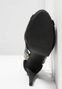 Chaussure unique noire à talon haut avec une semelle texturée et des brides à motif peau de serpent reposant contre un coin blanc.
