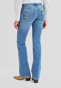 Moteris dėvi šviesiai mėlynas tiesias džinsines kelnes, baltą ilgomis rankovėmis marškinius ir smėlio spalvos kulniukus su aulinukais, stovinti nugara į žiūrovą.