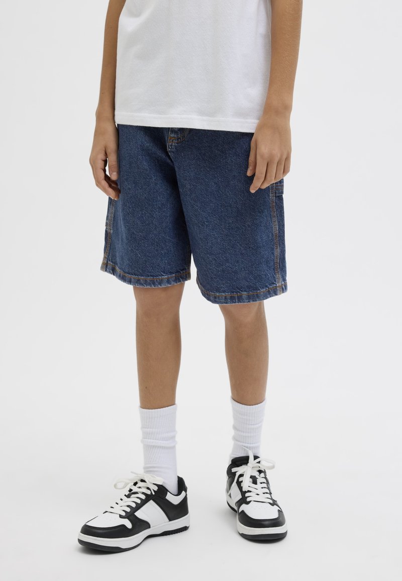 Bambino con maglietta bianca, pantaloncini di jeans blu, calzini bianchi e sneakers bianche e nere, in piedi su sfondo bianco.