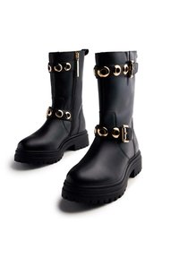 Botas de cuero negro hasta el tobillo con suela gruesa, que presentan acentos de herrajes circulares dorados y un cierre de cremallera lateral.