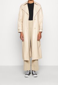 Beige lång kappa i blankt material med bred krage och dubbelknäppta knappar, kombinerad med beige randiga byxor och svarta sneakers.