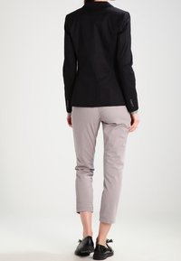 Osoba ubrana w czarną marynarkę, jasnoszare spódnice o kroju cropped oraz czarne mokasyny, stoi tyłem na tle gładkiego tła.