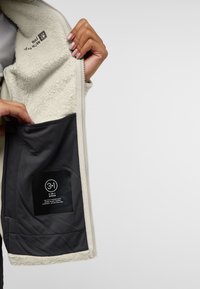 Fleecejack in lichtbeige met een ritssluiting en een donker binnenzakje met een zwart label dat een 3-in-1 systeem aangeeft.