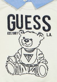 Λευκό πουκάμισο από ύφασμα με μπλε γιακά. Στην μπροστινή πλευρά υπάρχει κεντημένη αρκούδα π ου φοράει μπλούζα Guess με το λογότυπο.
