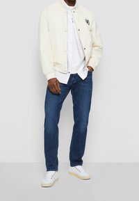 Chaqueta bomber crema clara con logotipo bordado, camisa blanca de botones, jeans azul oscuro y zapatillas blancas con suelas claras.