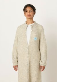 Dolg krem pulover s teksturirano pletenino, ki ima ovratnik, zapenjanje z gumbi in majhno modro oznako blagovne znamke na levi strani.