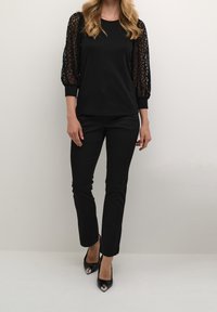 Schwarzes Oberteil mit Spitzen-Puffärmeln, kombiniert mit schwarzen, figurbetonten Hosen und spitzen High Heels. Glattes Material und strukturiertes Ärmel-Design.