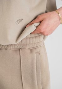 Beige színű pulóver, puha tapintású, mellkason logóval és kenguru zsebbel, bézs színű, hozzá illő melegítőnadrággal párosítva.