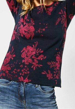 Frau trägt ein marineblaues Langarmshirt mit rotem Blumenmuster und blaue Jeans, mit erhobenen Armen über dem Kopf.