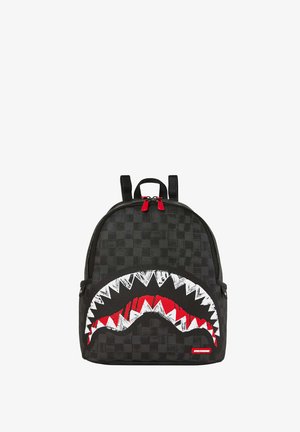 Sac à dos noir avec un motif à damier, arborant une grande bouche de requin brodée en blanc et rouge. Comprend deux tirettes de fermeture éclair rouges.