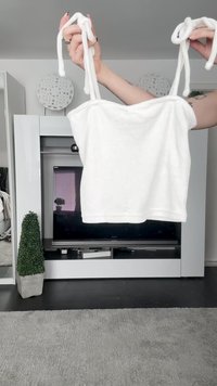 Top branco de alças tubulares, com uma textura suave e um design justo, apresentado contra um fundo de mobiliário e decoração.