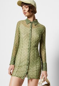 Vestido transparente abotoado em tons de verde, apresentando um padrão geométrico, colarinho e mangas longas. Tecido texturizado com uma silhueta ajustada.