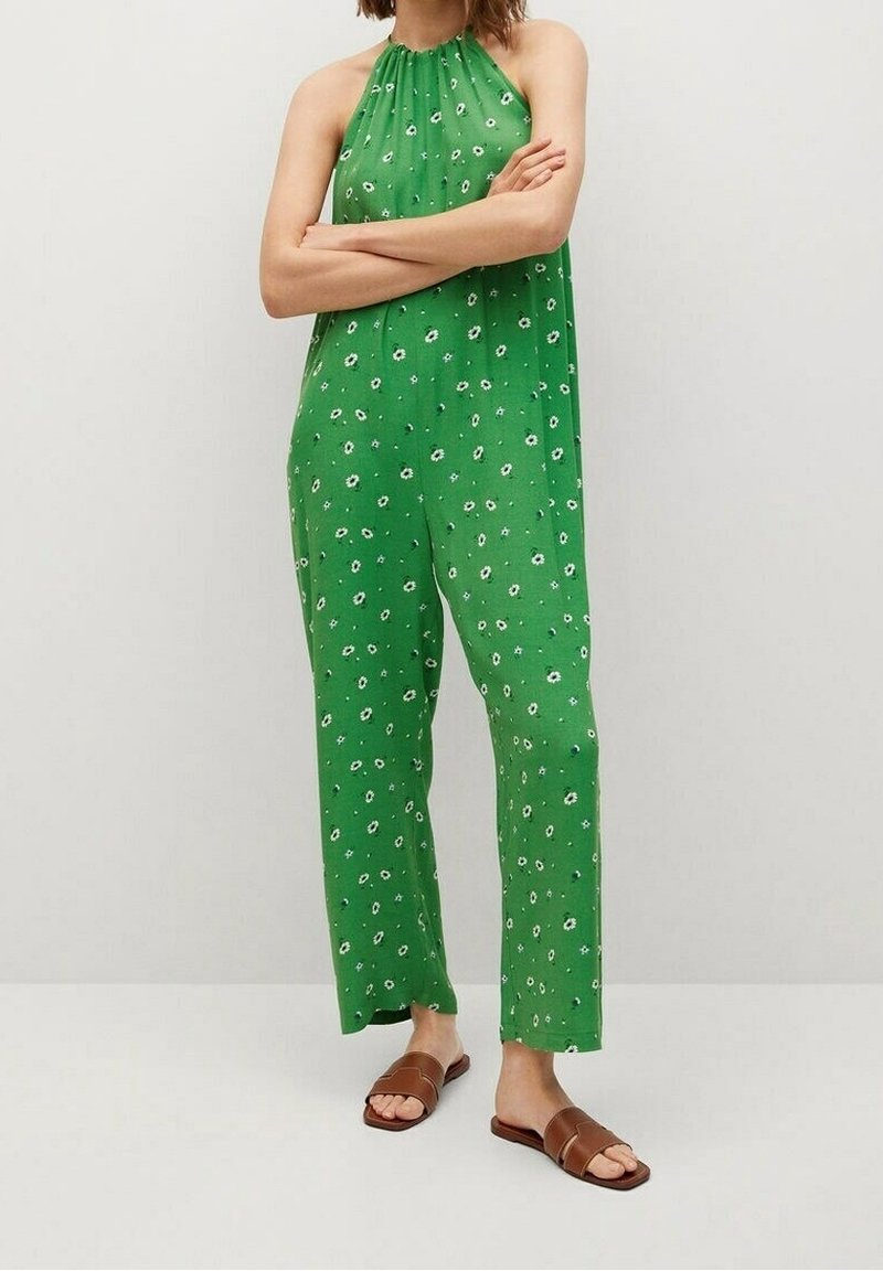 Femme debout, bras croisés, portant une combinaison verte à motifs floraux avec un col halter et des sandales plates marron sur un fond uni.