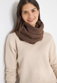 Écharpe en tricot marron avec un motif texturé, portée légèrement autour du cou, associée à un pull beige clair.