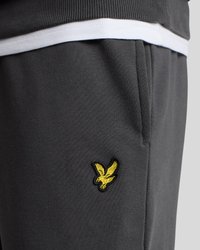 Pilkos sportinės kelnės su minkšta tekstūra, šoninėmis kišenėmis ir geltonu išsiuvinėtu erelio logotipu ant kairiojo kojos.