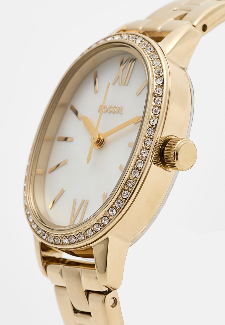 Montre dorée avec un bracelet en métal lisse, cadran blanc, index horaires dorés et accents en cristal autour de la lunette. Forme ronde classique.