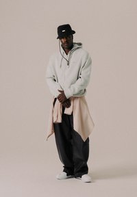 Hellgrauer Reißverschluss-Hoodie mit Kordelzug, getragen über einem pfirsichfarbenen Hemd mit Knöpfen; schwarze, weite Hose und weiße Schuhe; schwarzer Bucket Hat.