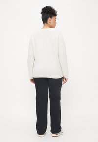 Lichtbeige, oversized trui met een ronde hals en verlaagde schouders, gedragen met donkere broek en meerkleurige sneakers.