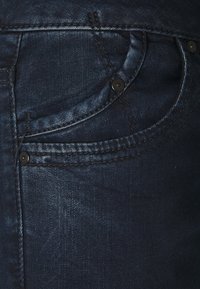 Mörkblå denimjeans med en framficka i rundad form, synliga sömmar och svarta metalldetaljer för stängning.