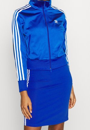 Person trägt eine blaue Adidas Trainingsjacke und passenden Rock mit weißen Streifen an Ärmeln und Rockseiten, steht vor einem schlichten Hintergrund.