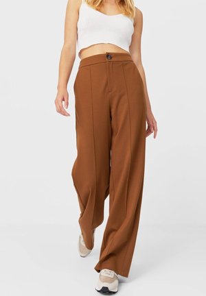 Mujer con pantalones marrones de tiro alto y pierna ancha con botón delantero, top corto blanco y zapatillas beige caminando hacia adelante.
