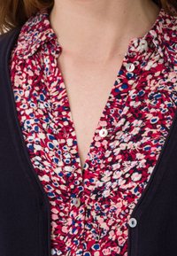 Gros plan d'une personne portant un chemisier rouge à fleurs avec des boutons blancs et un cardigan bleu marine foncé par-dessus.