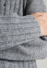 Grå stickad tröja med ribbad textur, med synliga stygn och en snug passform runt ärmarna. Tillverkad i ett mjukt, varmt material.
