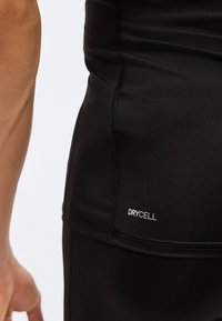 Κοντινό πλάνο στο πλευρό του κορμού ενός ατόμου που φοράει μαύρο αθλητικό μπλουζάκι με το λογότυπο "DRYCELL" κοντά στο τελείωμα.