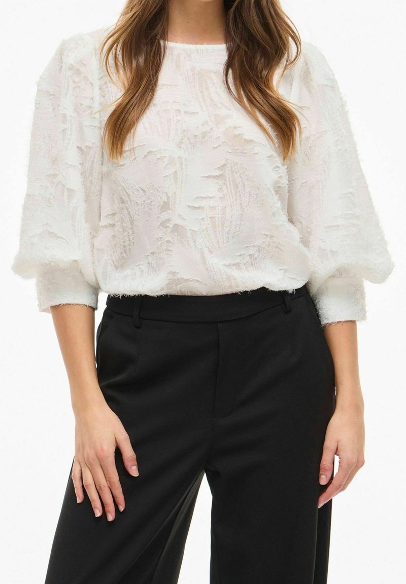 Femme portant un chemisier blanc texturé avec des manches ballon, associé à un pantalon noir taille haute, debout les mains détendues le long du corps.