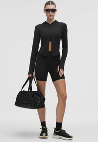 Jachetă de athleisure neagră, cu fermoar, de croială scurtă, asortată cu pantaloni scurți negri. Modelul ține o geantă de voiaj neagră. Tenisi sport negri completează look-ul.
