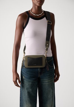 Groene crossbody tas met een gestructureerde zwarte voorzak, gouden hardware en een verstelbare schouderriem; gedragen boven een lichte tanktop en denim jeans.