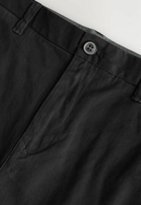 Zwarte broek met een tailleband met een knoop, riemlussen en zichtbare stiksels van de voorzak op een lichtgrijze achtergrond.