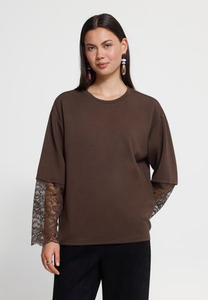 Žena s dlhými tmavými vlasmi nosiaca hnedý oversized top vrstvený cez hnedé čipkové rukávy a čierne nohavice, stojaca pred jednoduchým bielym pozadím.
