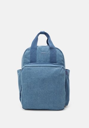 Sac à dos en tissu denim bleu avec deux poignées, une poche zippée à l'avant et des poches latérales, présenté sur fond blanc.