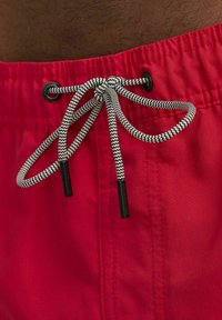 Los bañadores rojos cuentan con un cordón ajustable con un patrón en zigzag blanco y negro, asegurado por ojetes negros. Tejido liso y ligero.
